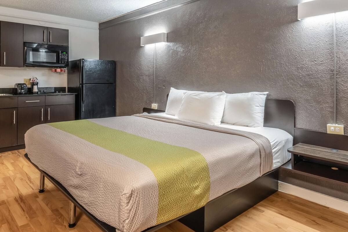 Un dormitorio moderno con cama, mesitas de noche, televisor integrado y una kitchenette con microondas y refrigerador.