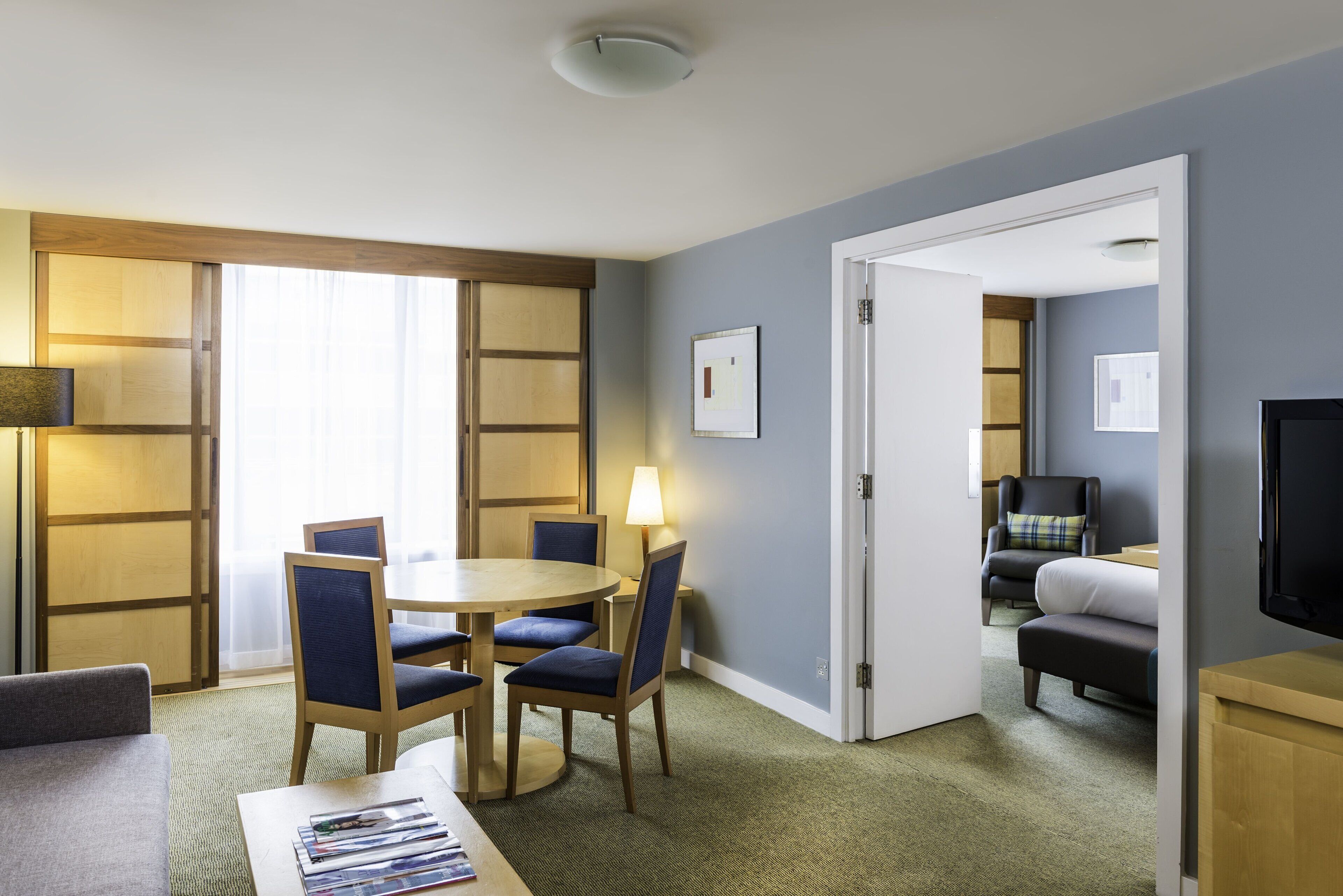 Habitación de hotel con comedor, cama, televisor y puerta que da a otra habitación.