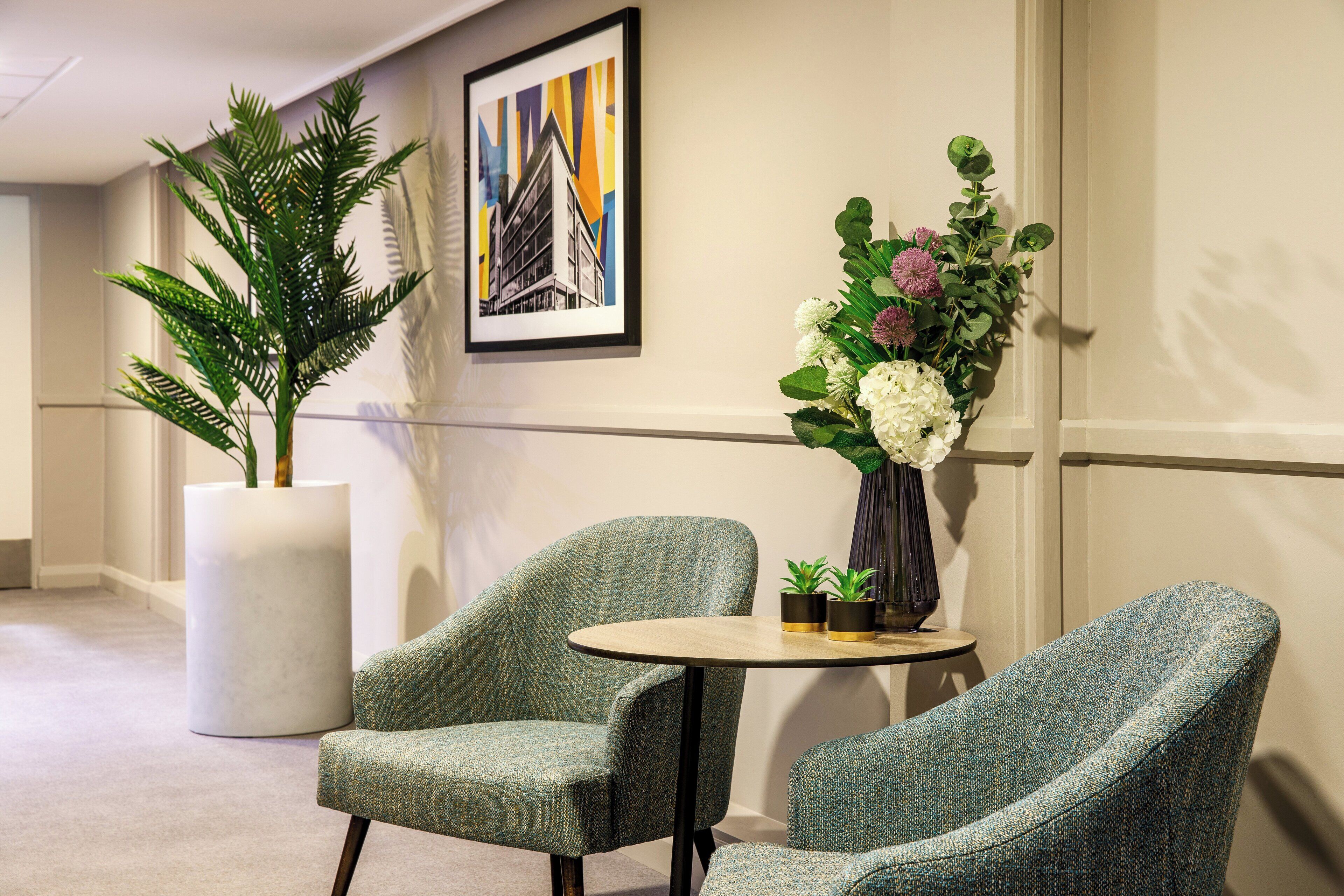 A lobby with two green upholstered chairs, a round table, a large potted plant, and a framed artwork on the wall.