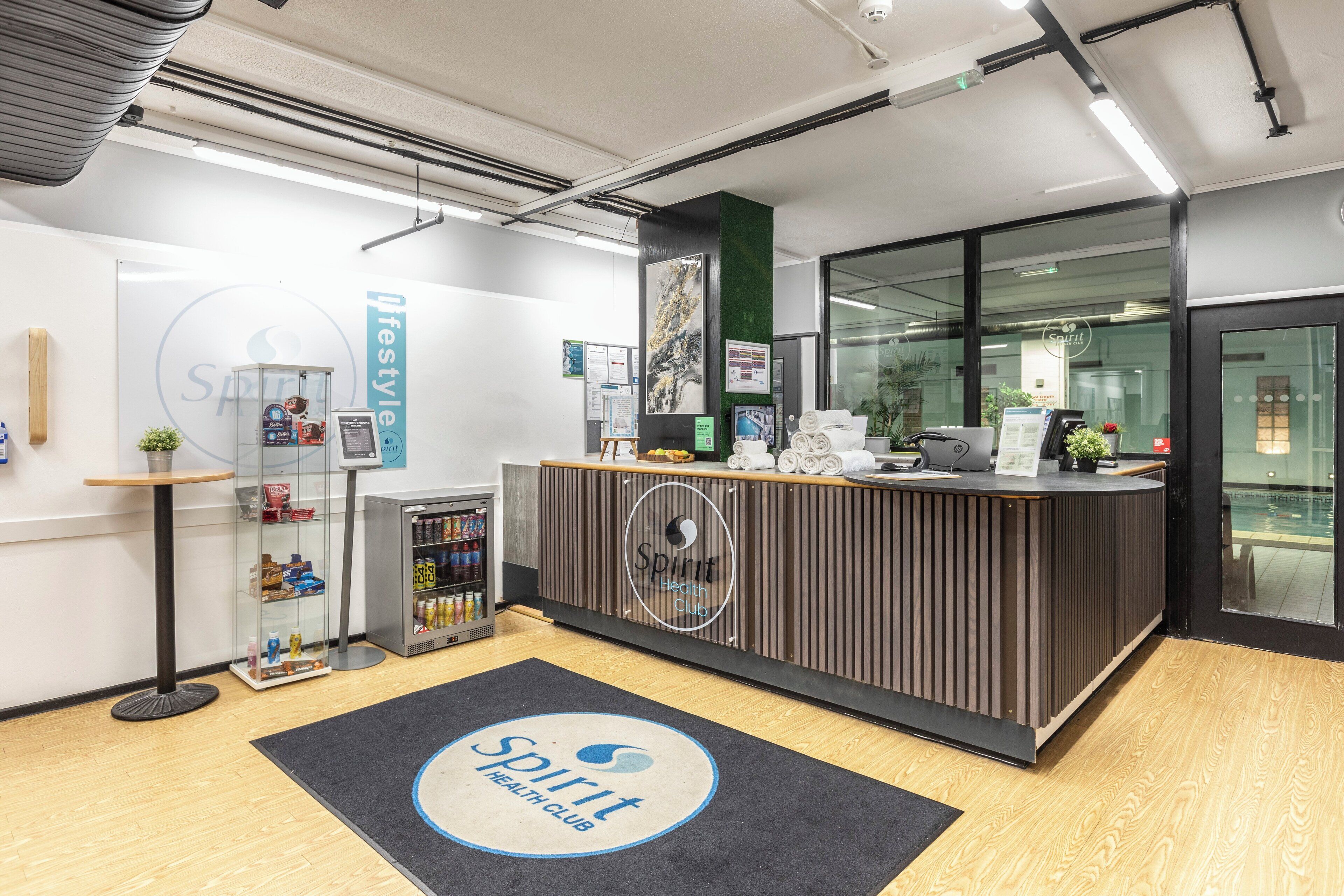 A reception area with a wooden reception desk, a glass display case, and a black mat with the logo 'Spirit Health Club'.