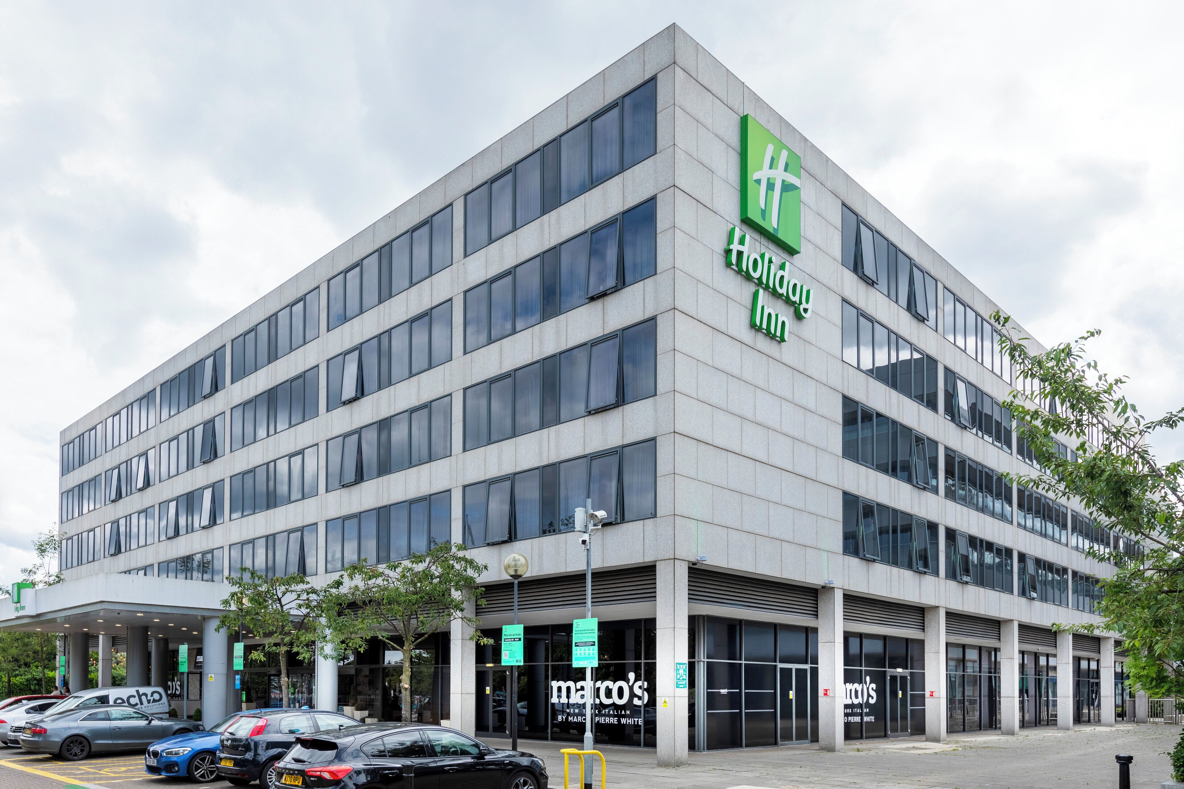 A modern Holiday Inn hotel with a large sign, multiple floors, and a parking lot with several cars.