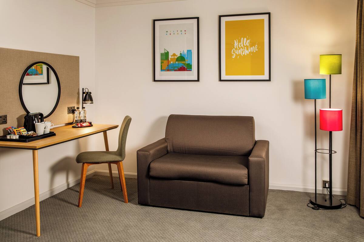 A hotel room with a brown sofa, a wooden desk with a coffee maker, a chair, and a floor lamp with multiple colored shades.