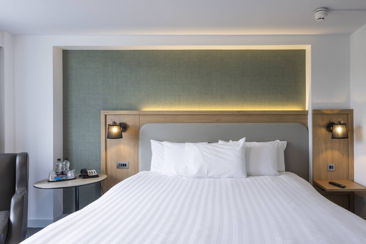 A hotel room with a large bed, two bedside tables, a small round table with a chair, and a wall-mounted lamp.