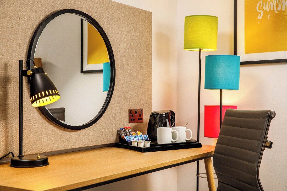 A hotel room with a round mirror, a desk with a coffee maker, cups, and a tray of toiletries.