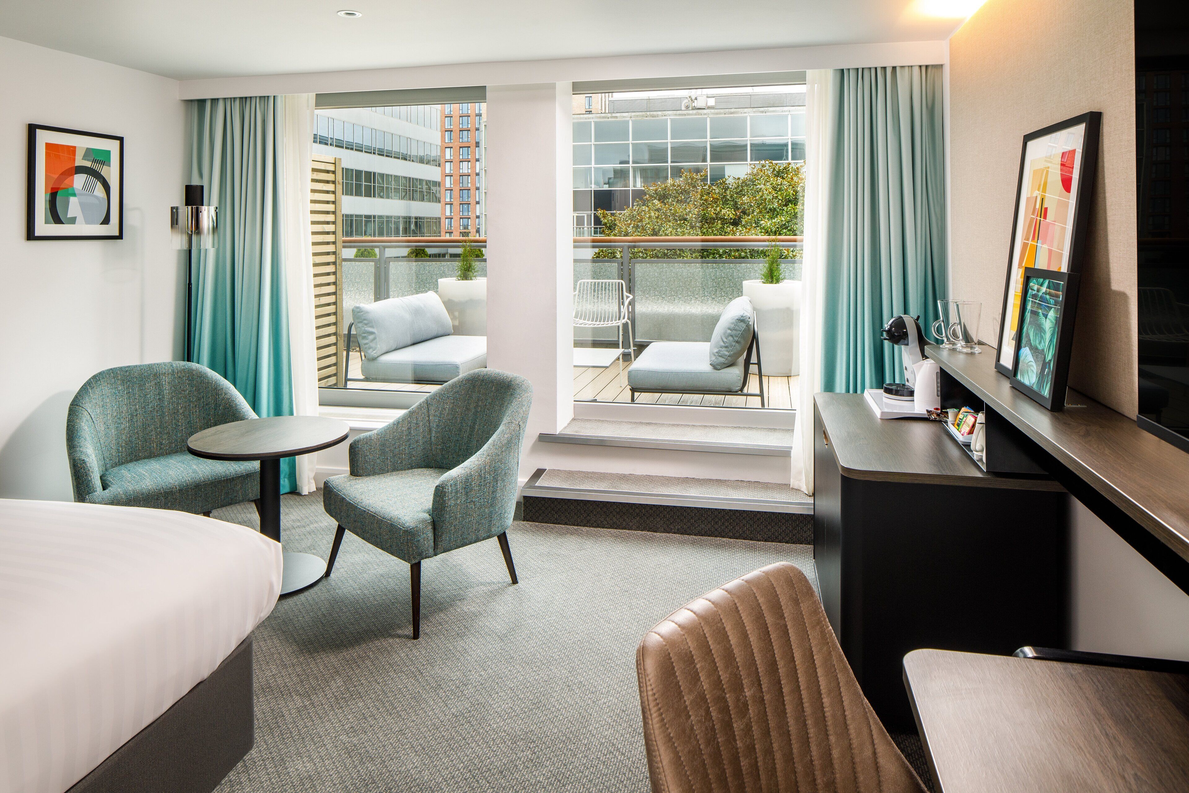 A hotel room with a bed, two armchairs, a small table, a desk, and a balcony with a view of buildings and greenery.