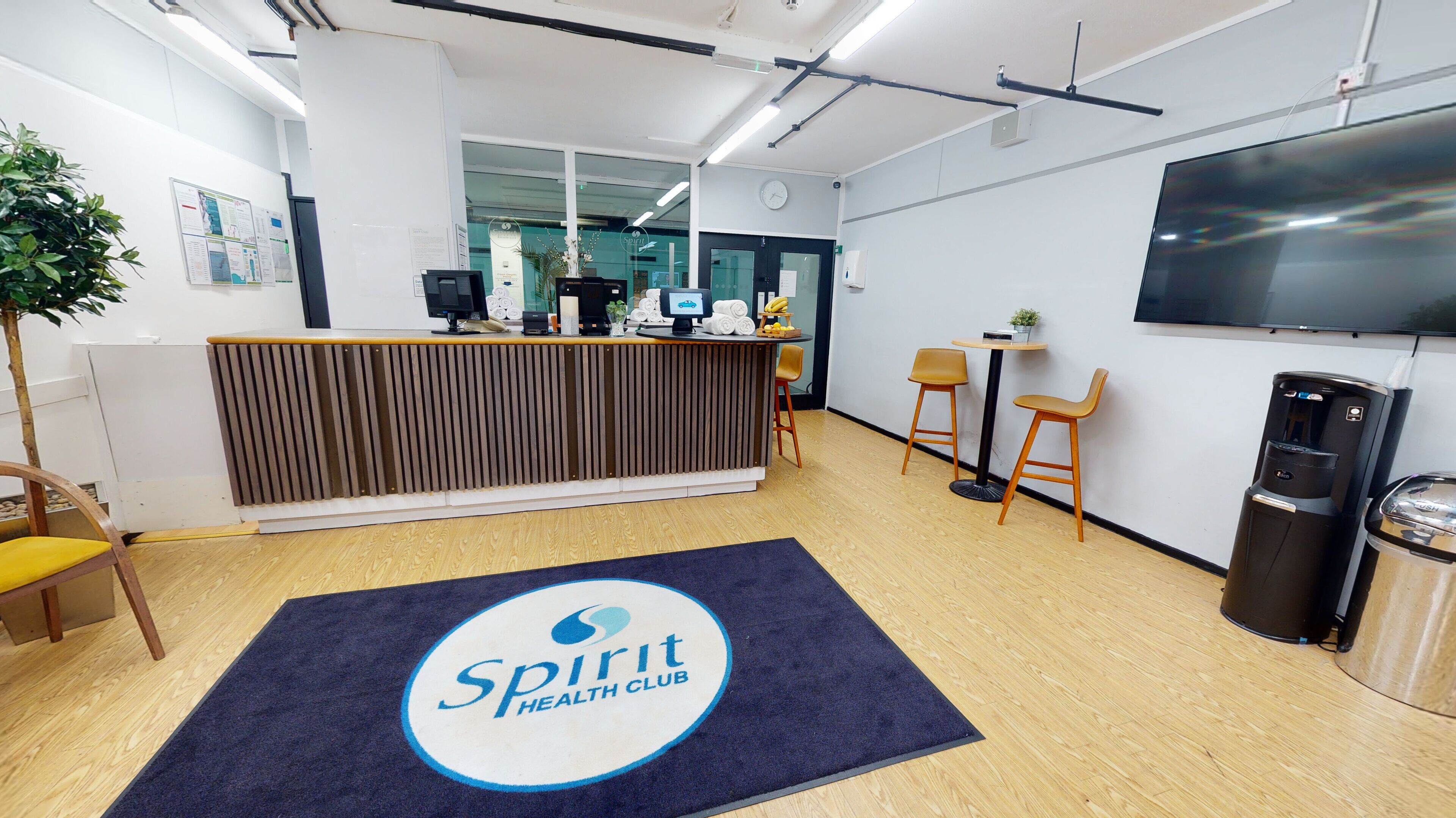 Un área de recepción con un mostrador de madera, una alfombra azul con el logotipo de 'Spirit Health Club', dos sillas de madera, una mesa pequeña con una planta y un televisor montado en la pared.