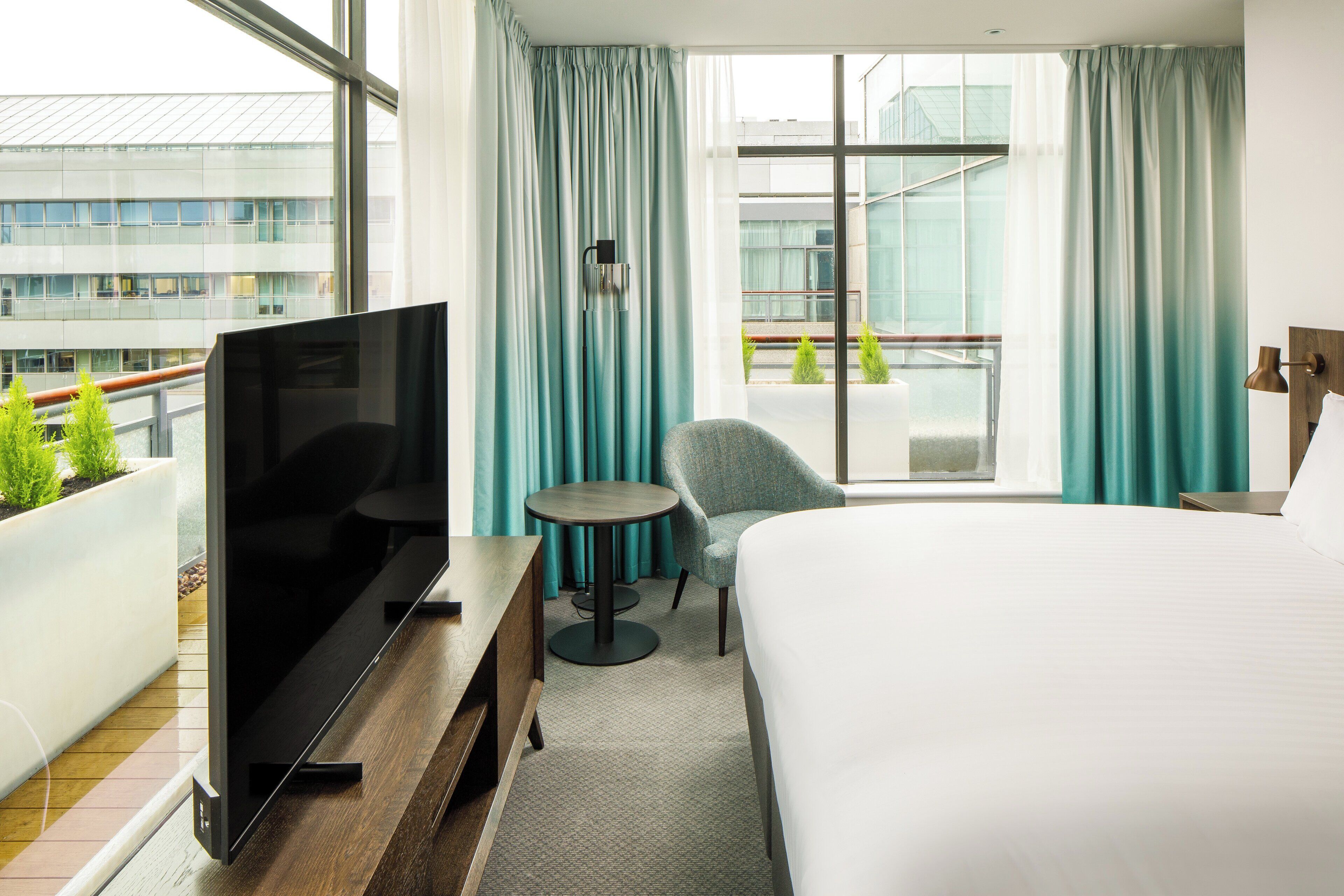 A hotel room with a bed, a TV, a small table, a chair, and a balcony with a view of buildings and greenery.