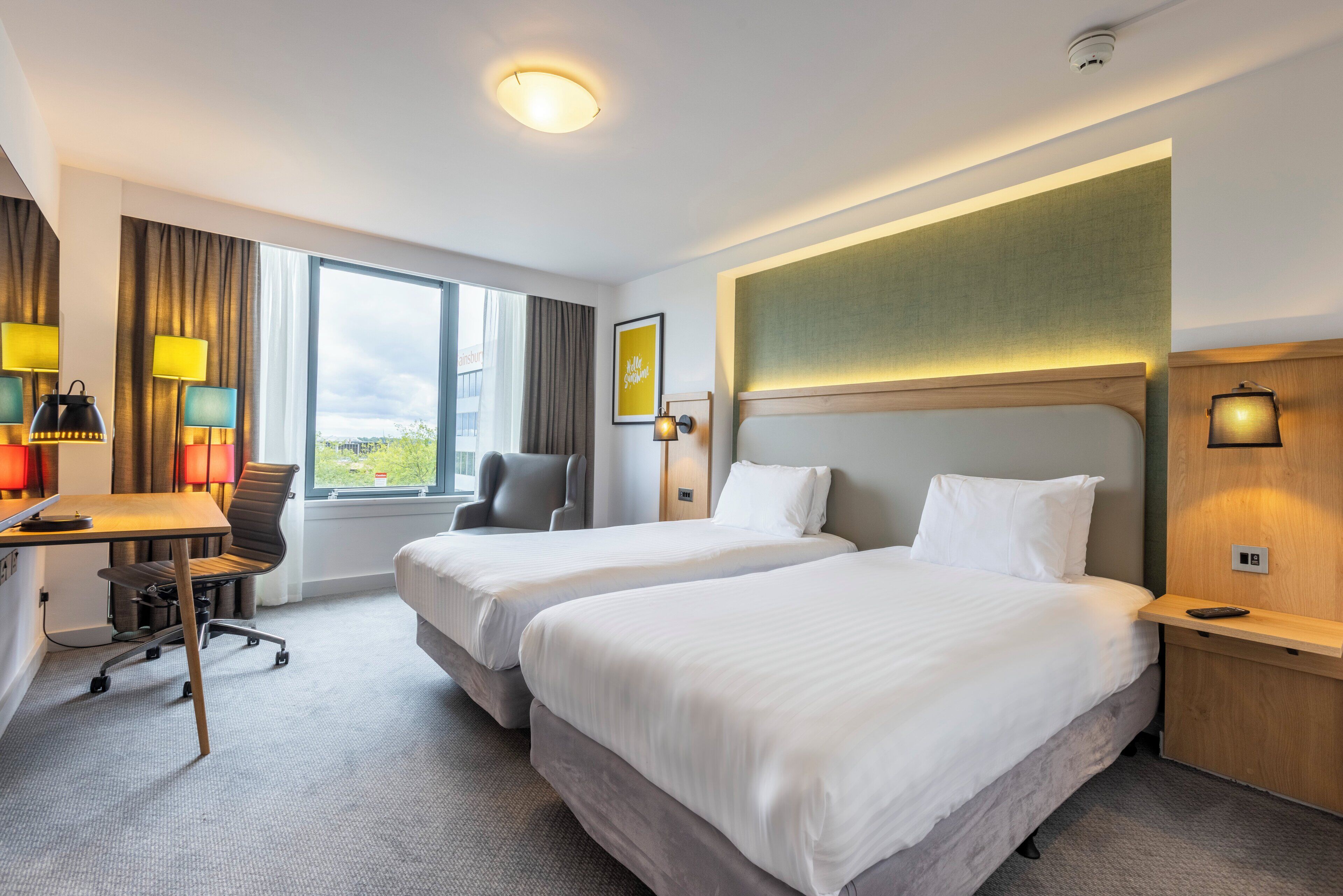 Ein Hotelzimmer mit zwei Betten, einem Schreibtisch und einem Sessel. Ein Fenster bietet einen Blick ins Freie.