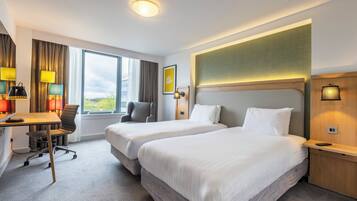Ein Hotelzimmer mit zwei Betten, einem Schreibtisch und einem Sessel. Ein Fenster bietet einen Blick ins Freie.