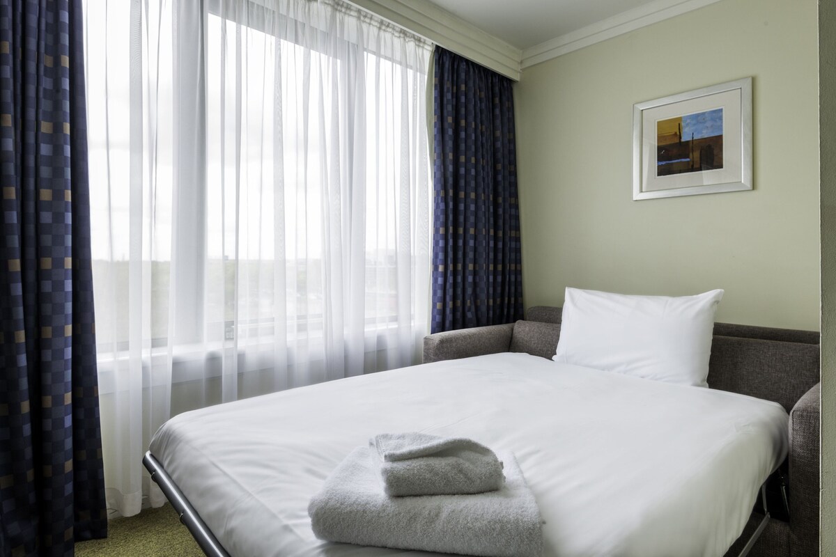 A hotel room with a bed, a chair, a pillow, a framed picture on the wall, and curtains.