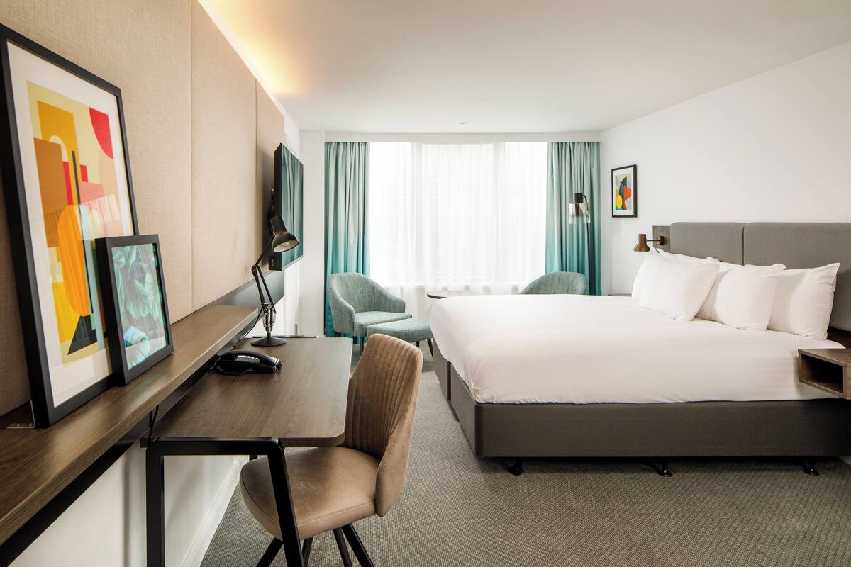 A hotel room with a bed, a desk, a chair, a TV, and a window with curtains.