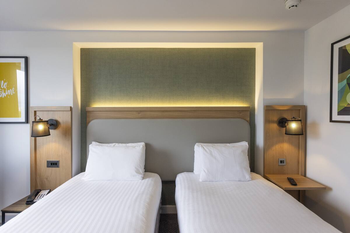 A hotel room with two beds, a headboard, bedside lamps, a phone, and a wall-mounted light fixture.