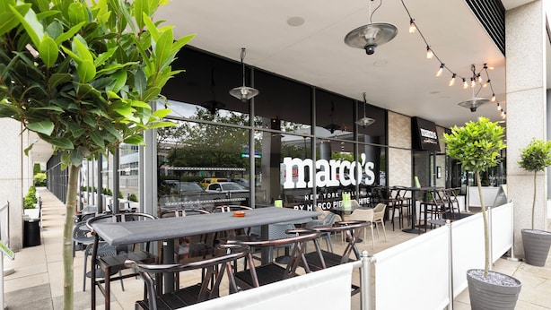 An outdoor seating area with tables and chairs, a large window with the name "marco's" visible, and potted plants. - Holiday Inn Milton Keynes - Central by IHG (Milton Keynes)