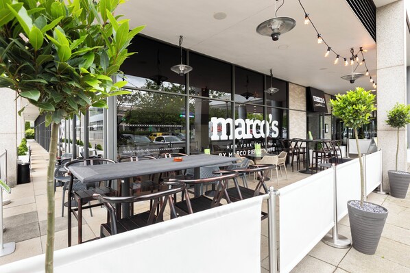 An outdoor seating area with tables and chairs, a large window with the name "marco's" visible, and potted plants. - Holiday Inn Milton Keynes - Central by IHG (Milton Keynes)