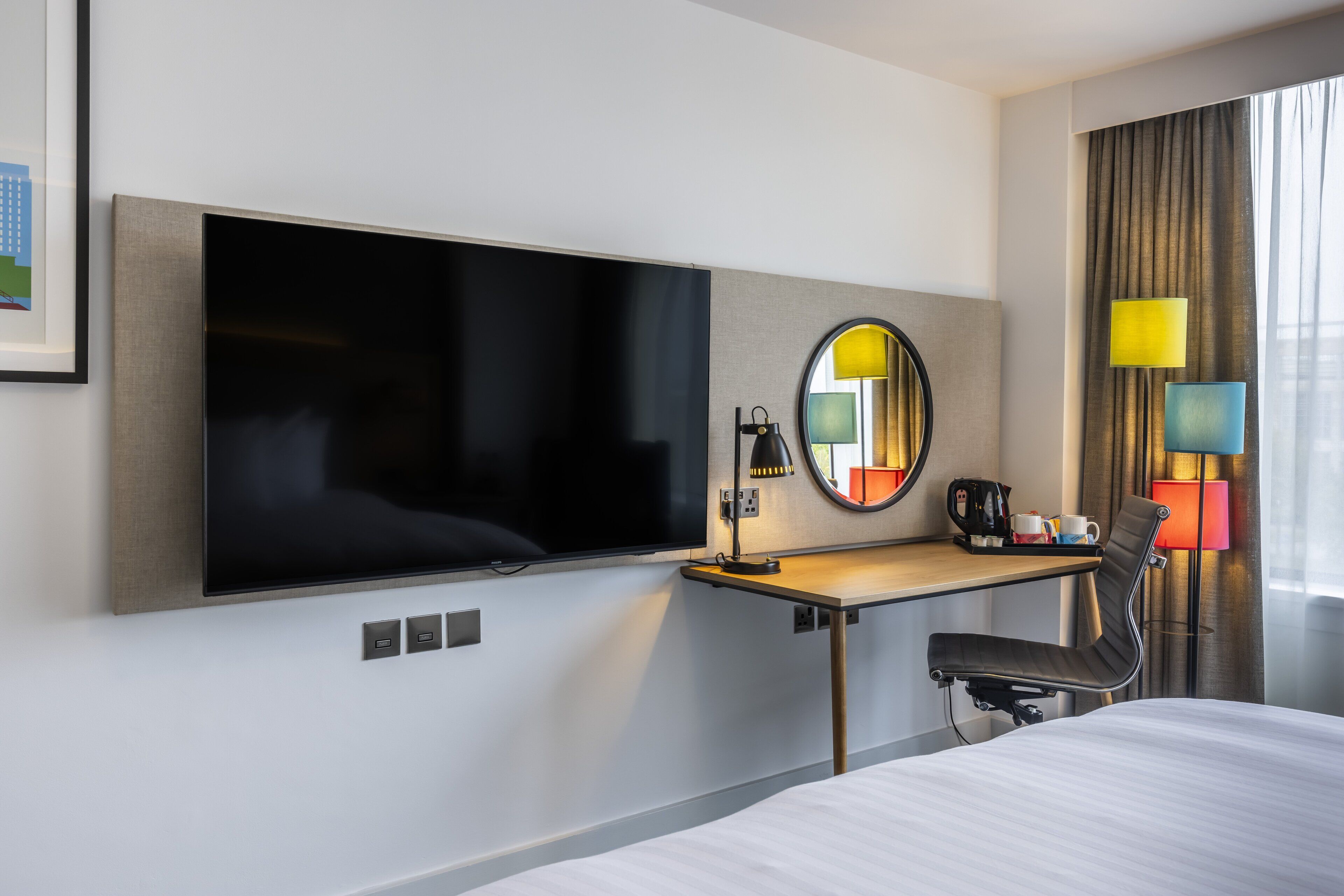 Habitación de hotel con televisor de pantalla plana montado en la pared, escritorio con silla, cama y una ventana grande con cortinas.