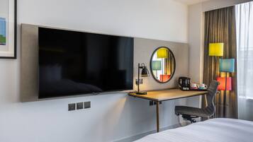 Habitación de hotel con televisor de pantalla plana montado en la pared, escritorio con silla, cama y una ventana grande con cortinas.