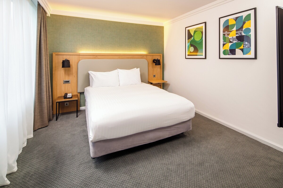 A hotel room with a large bed, a wooden headboard, a bedside table with a lamp, and two abstract paintings on the wall.