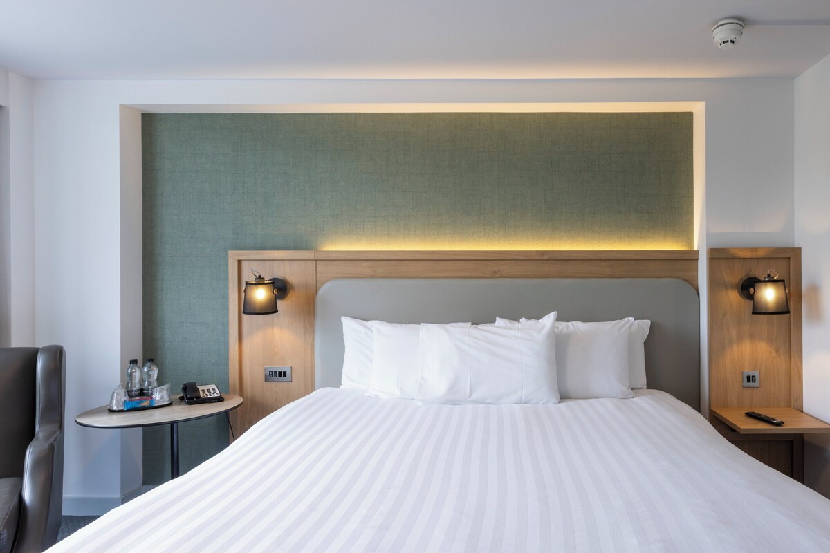A hotel room with a large bed, two bedside tables, a small round table with a chair, and a wall-mounted lamp.