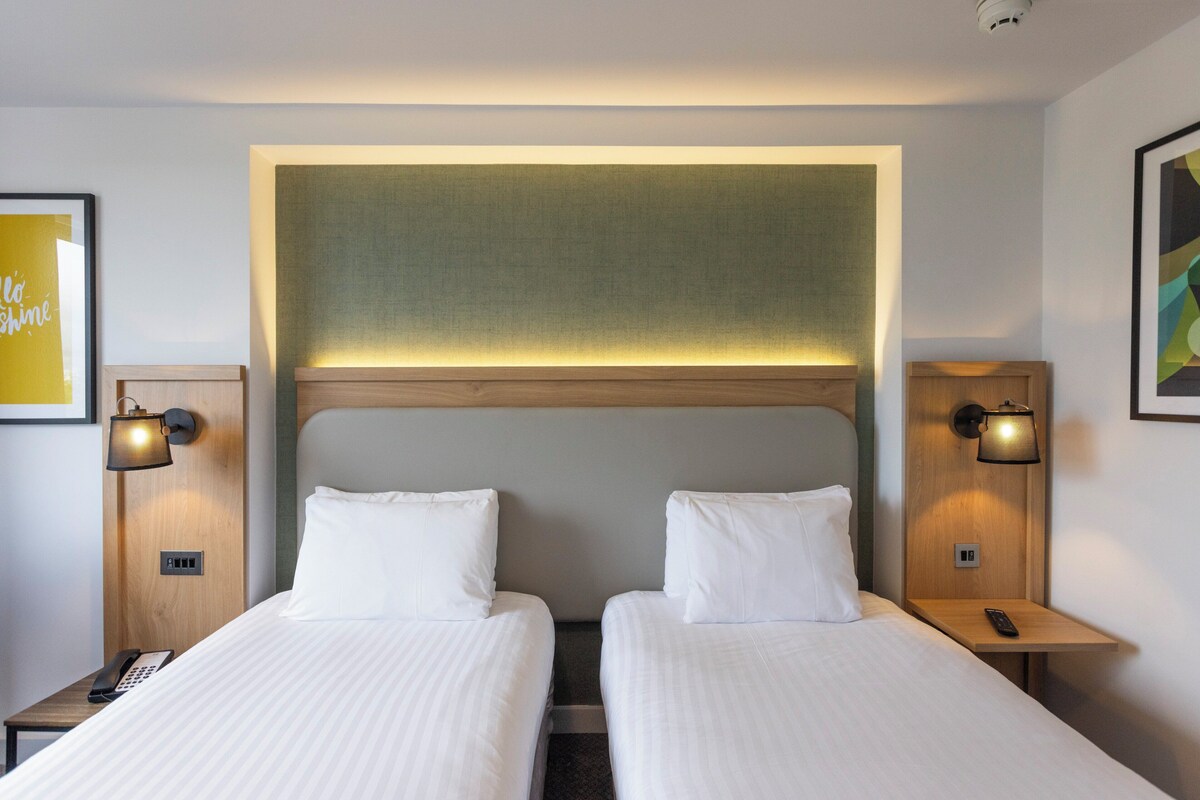 A hotel room with two beds, a headboard, bedside lamps, a phone, and a wall-mounted light fixture.