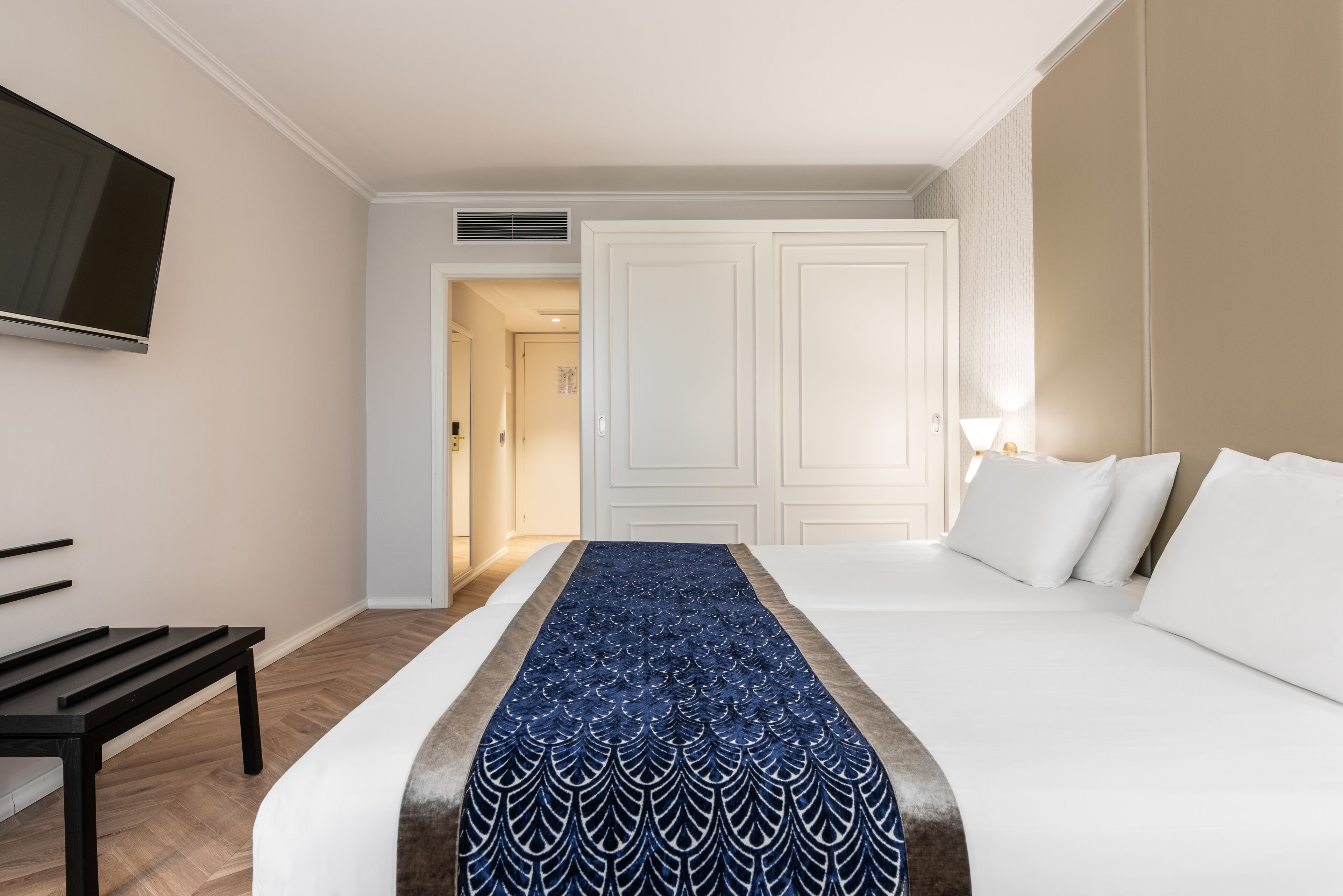 A hotel room with a bed, a bench, a TV, and a door leading to another room.