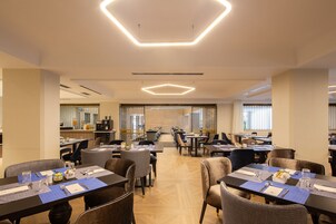 Un comedor moderno con lámparas de techo hexagonales, varias mesas con individuales azules y una variedad de sillas tapizadas.