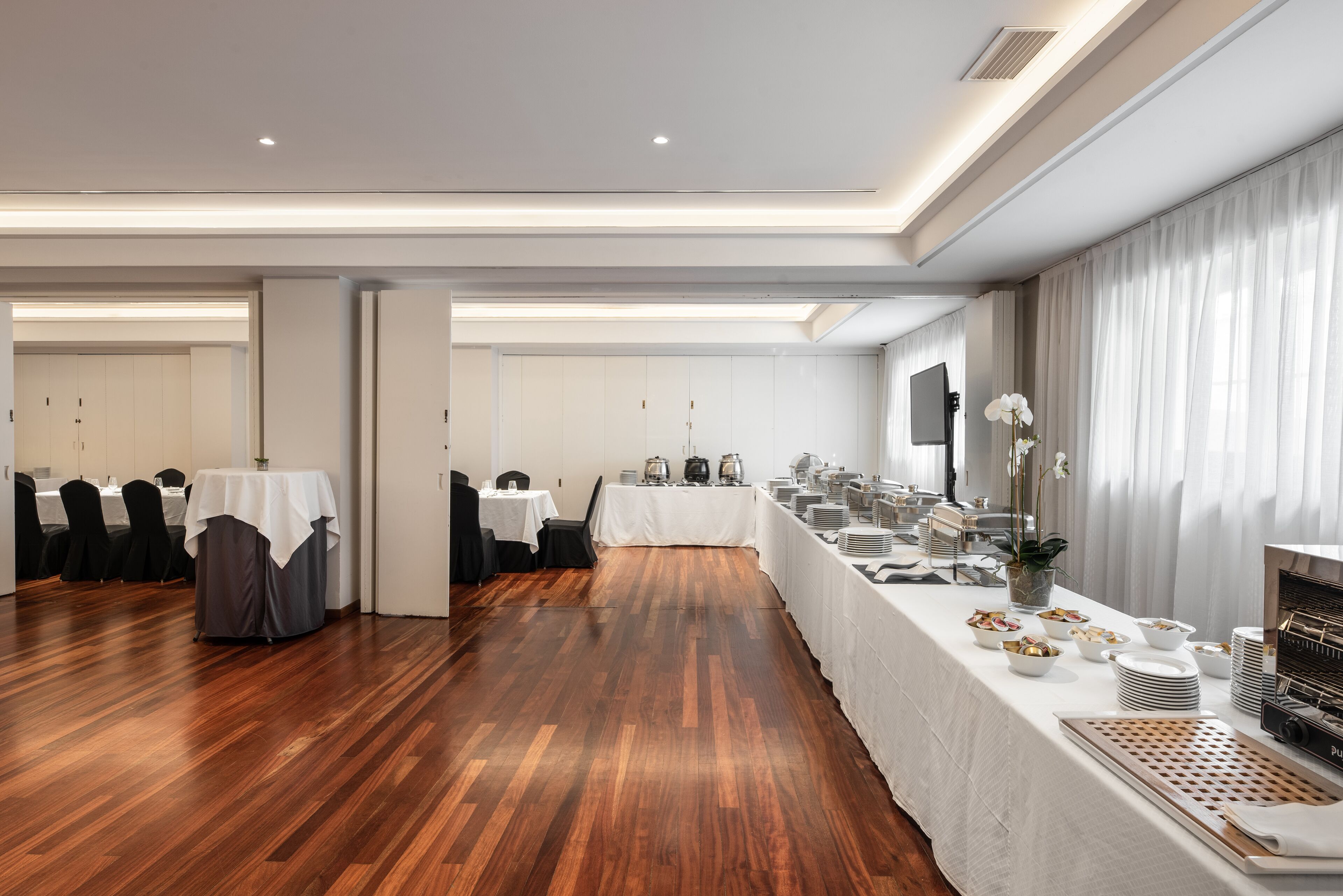 A spacious room with a long table set for a buffet, wooden flooring, and a modern ceiling design.
