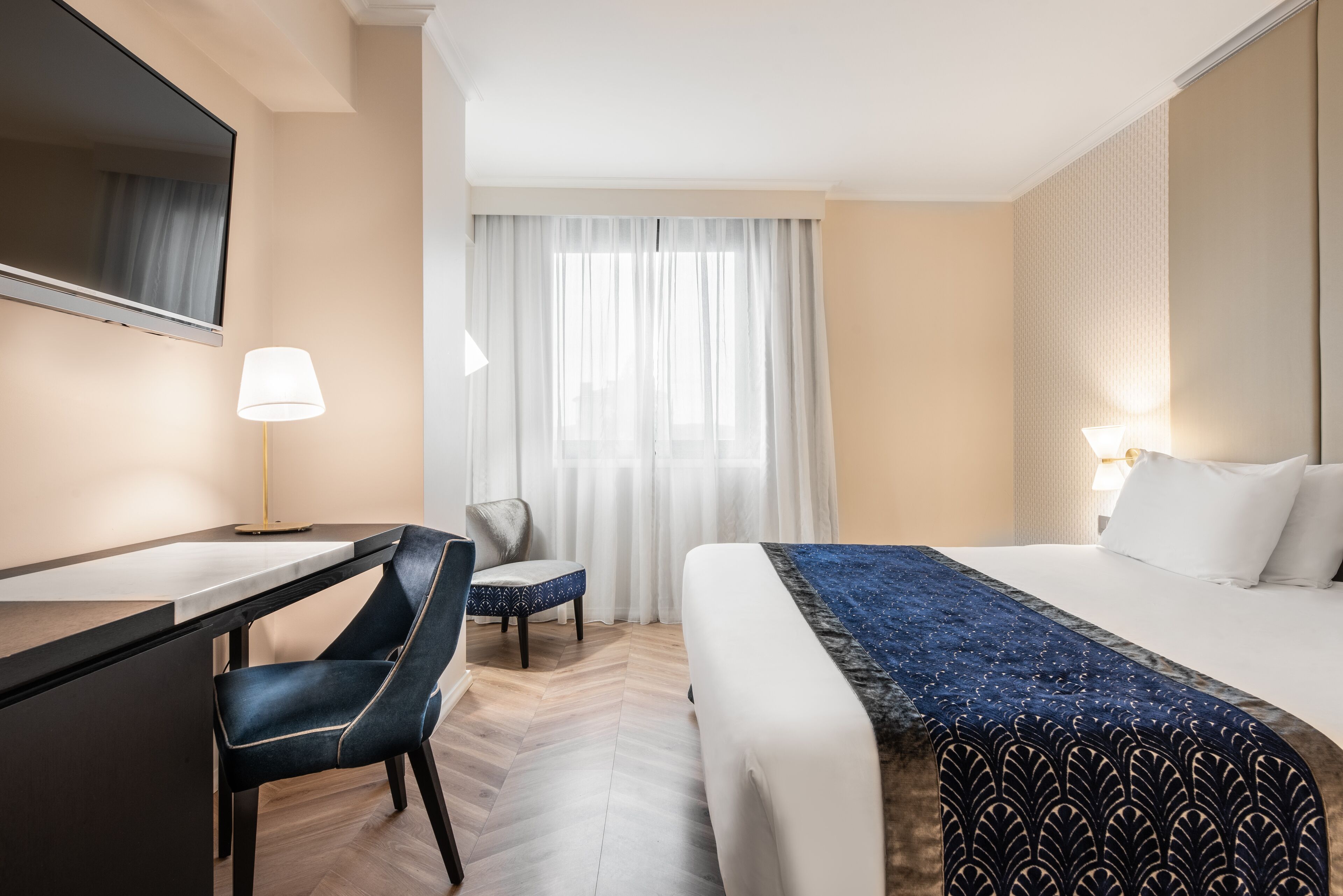 Une chambre d’hôtel comprenant un lit, un bureau, une chaise, une grande fenêtre avec des rideaux et une lampe.