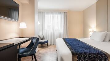 Ein Hotelzimmer mit einem Bett, einem Schreibtisch, einem Stuhl, einem großen Fenster mit Vorhängen und einer Lampe.