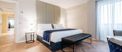 A modern hotel room with a large bed, a small bedside table, a bench, and a view of the hallway.
