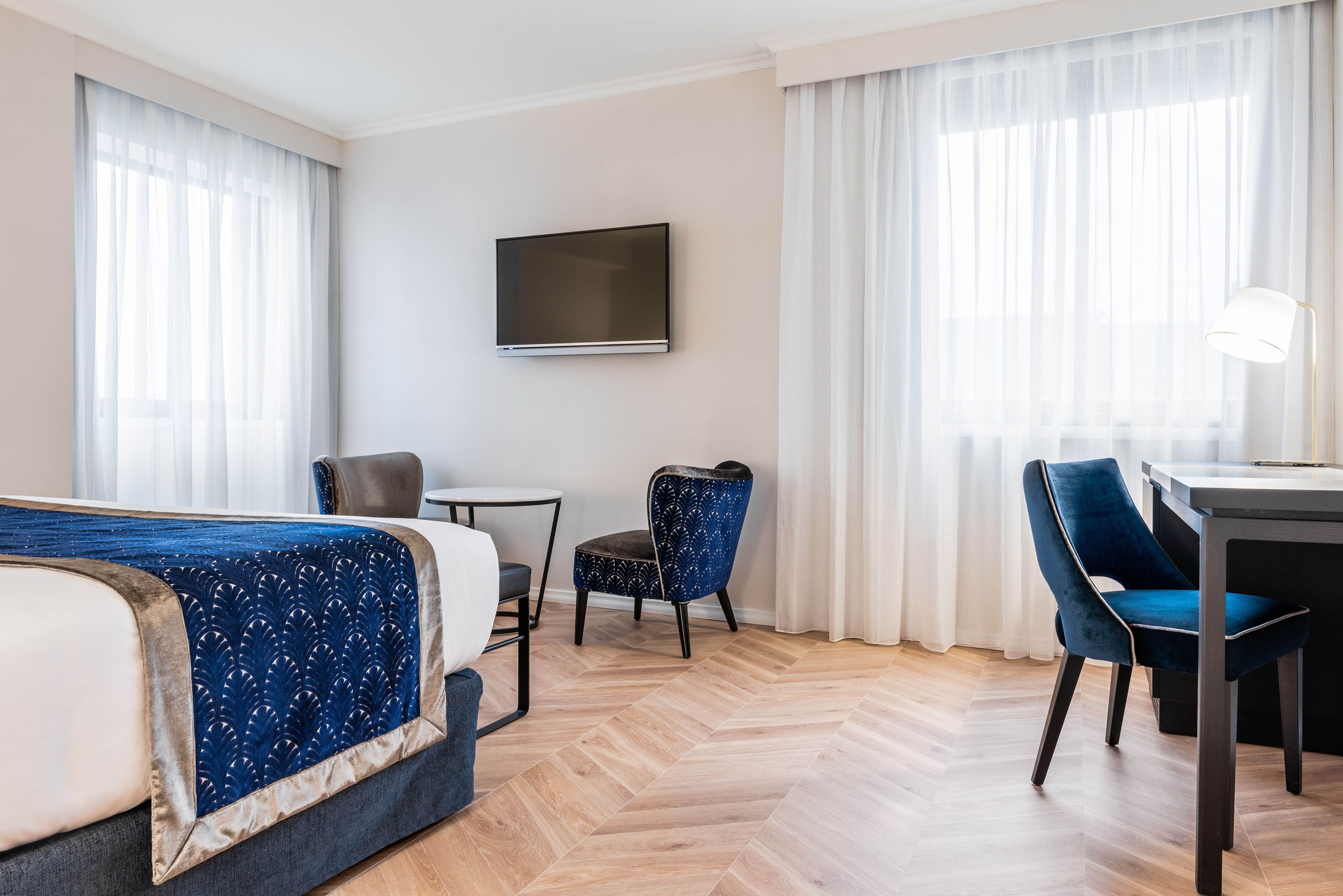 A hotel room with a bed, a desk, a chair, a TV, and large windows with curtains.