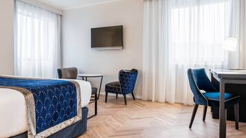 A hotel room with a bed, a desk, a chair, a TV, and large windows with curtains.