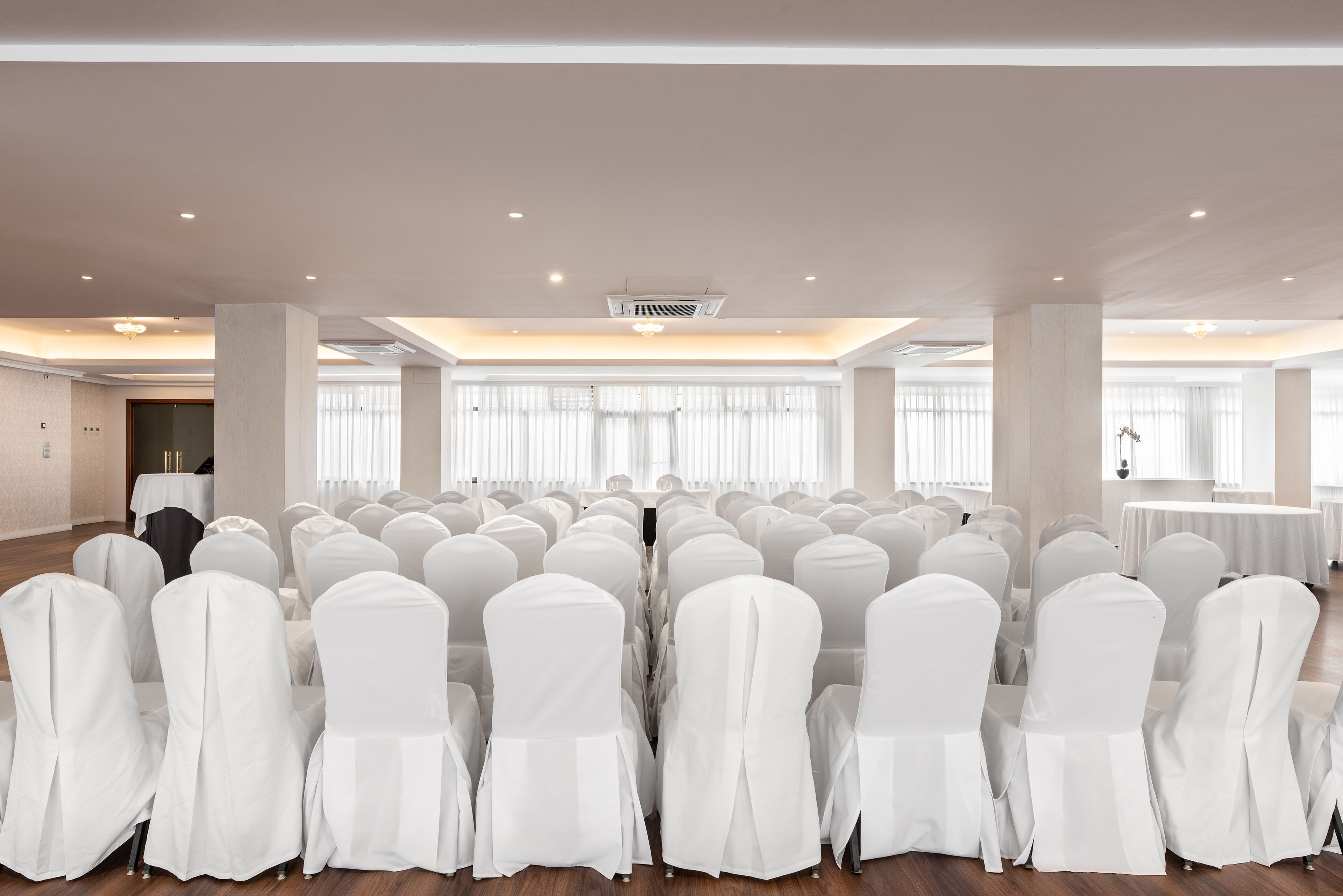 Un salón de conferencias con filas de sillas blancas, ventanas amplias y una mesa cubierta con un mantel blanco.