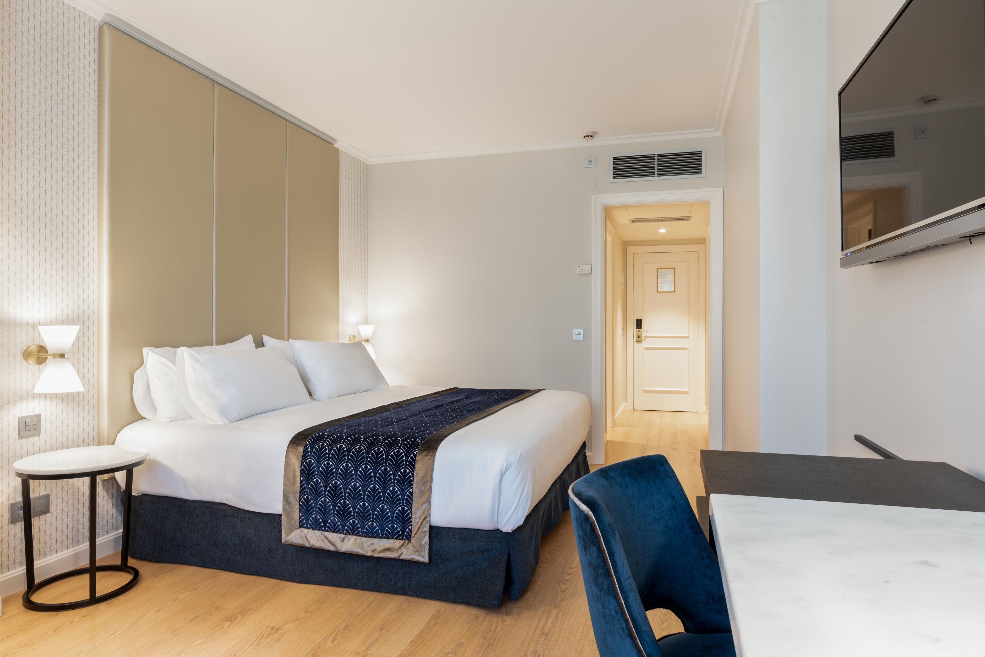 Una habitación de hotel con cama, mesita de noche, silla y televisor montado en la pared.