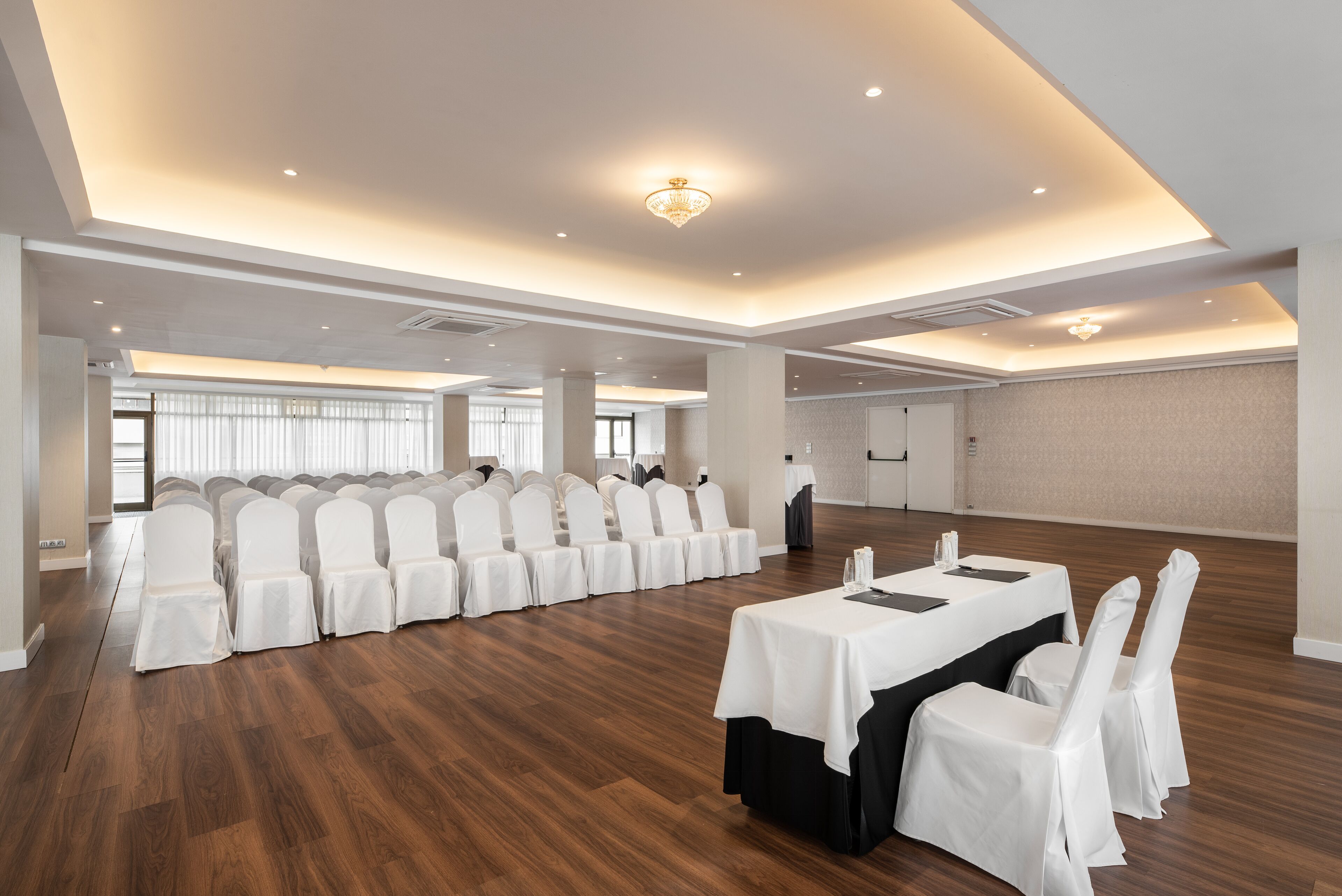 Een ruime congreszaal met rijen witte stoelen, een lange tafel en een kroonluchter.