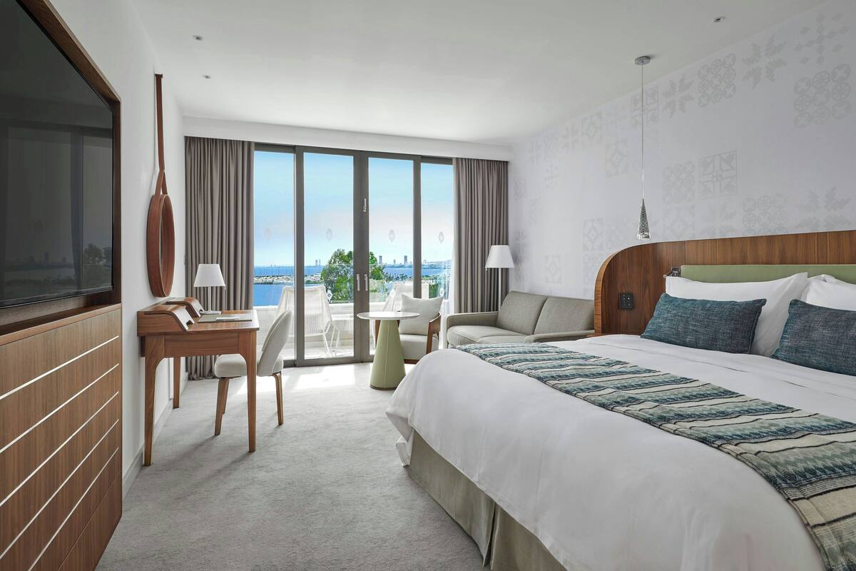 A hotel room with a large bed, a desk, a chair, a TV, and a view of the city through large windows.