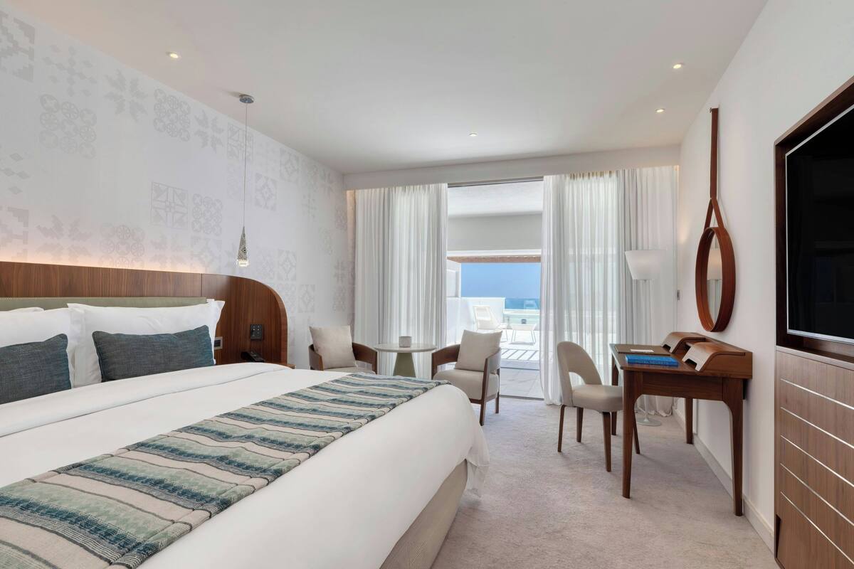 A hotel room with a large bed, a desk, a chair, a TV, and a balcony with curtains.