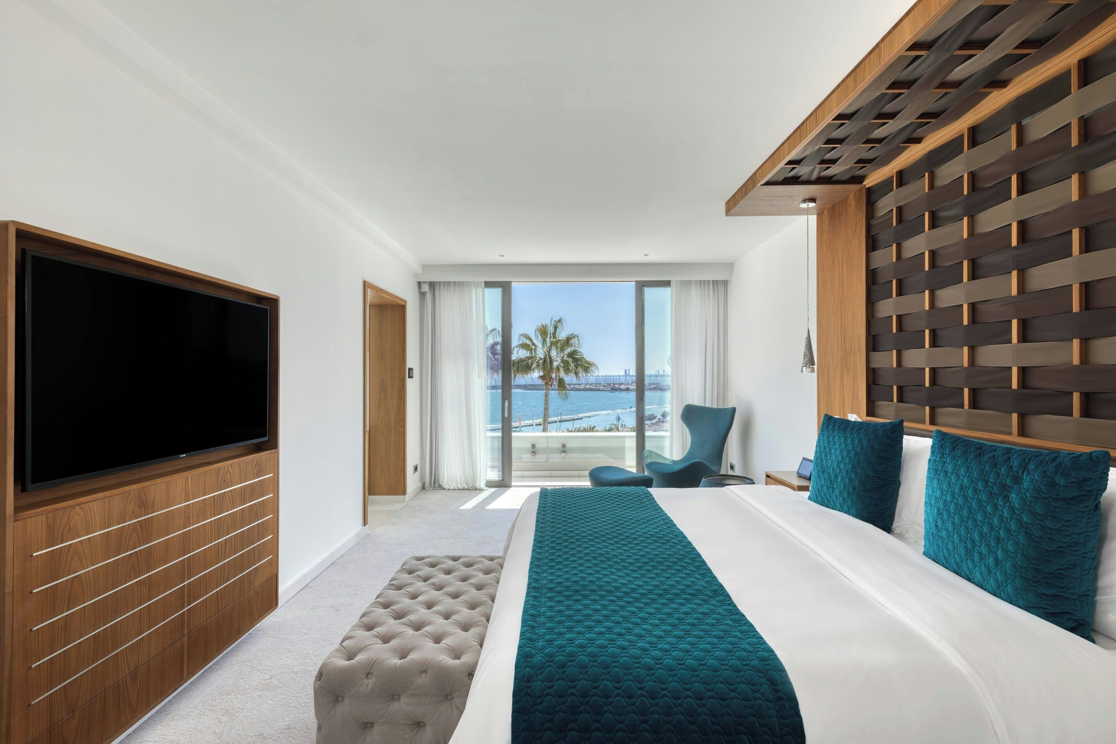 A hotel room with a large flat-screen TV, a bed with teal bedding, a wooden headboard, and a view of the ocean.