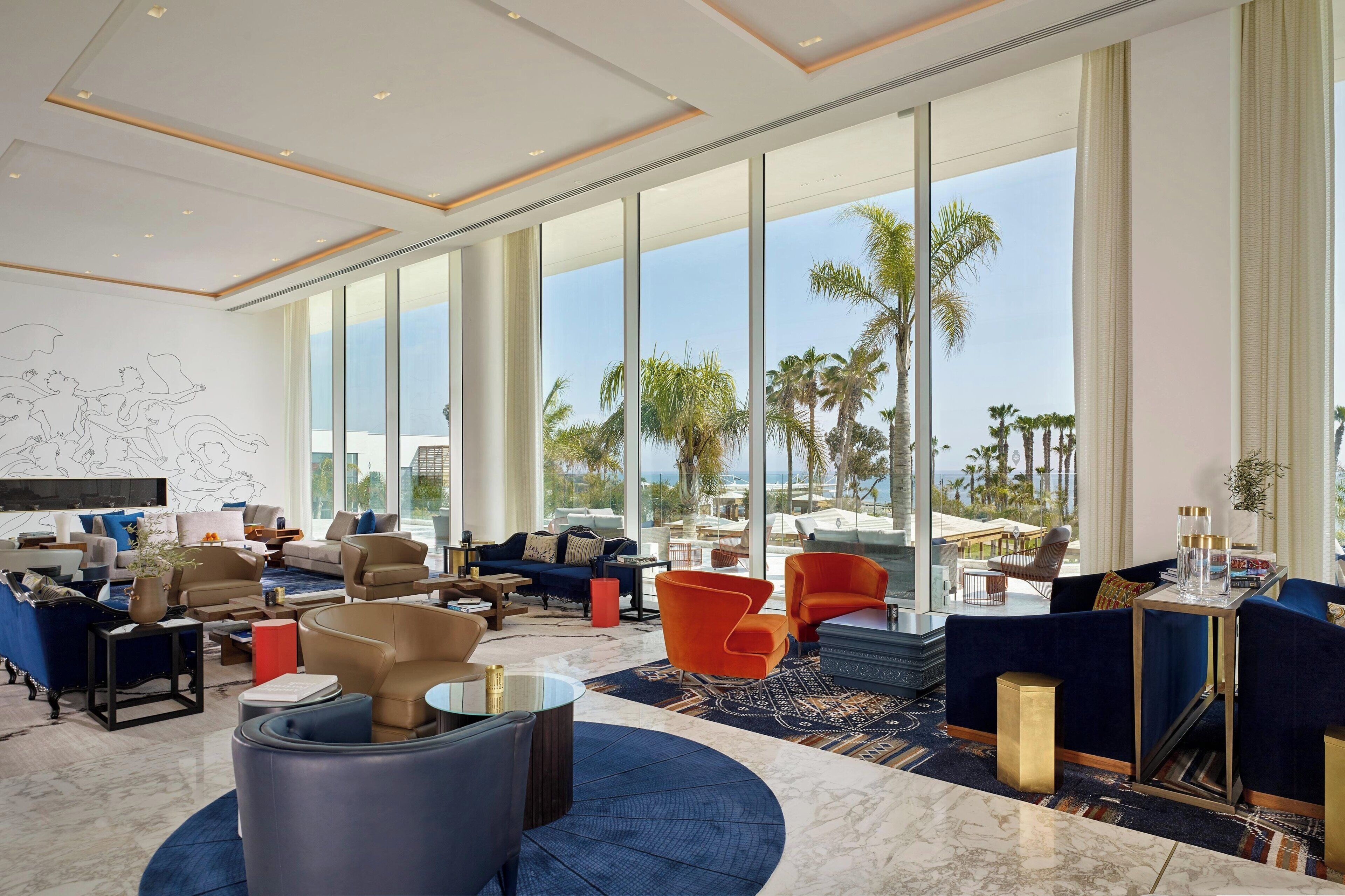 A spacious hotel lobby with a large seating area, a television, and large windows offering a view of palm trees and the ocean.