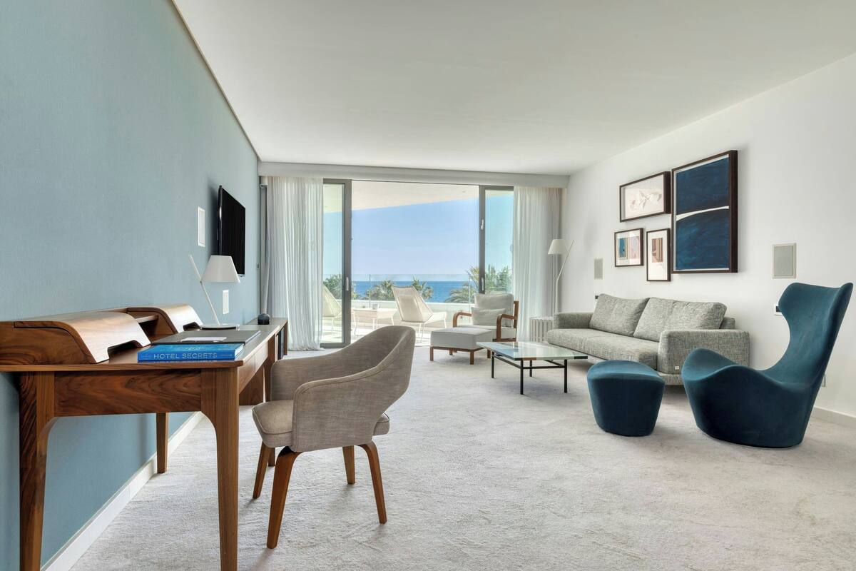 A modern hotel room with a desk, chairs, a sofa, a coffee table, and a view of the ocean.