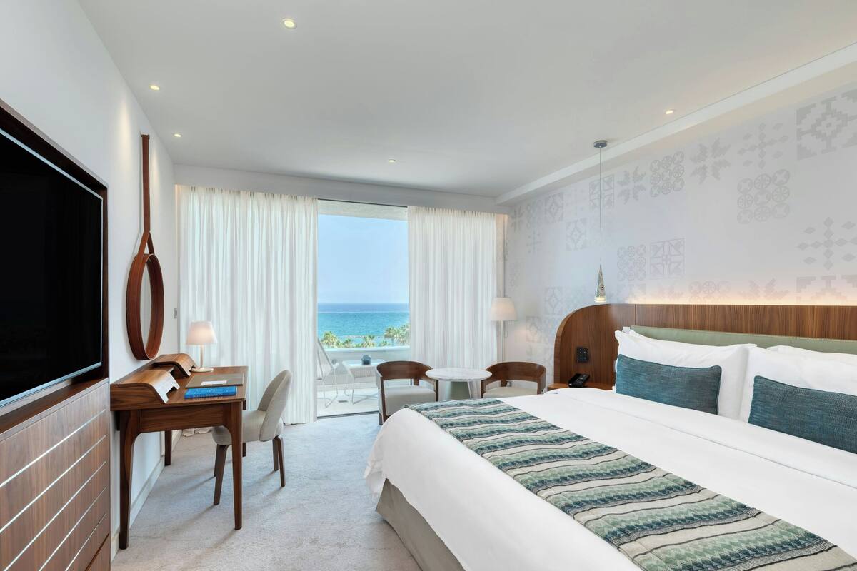 A hotel room with a large bed, a desk with a computer, a TV, and a view of the ocean.