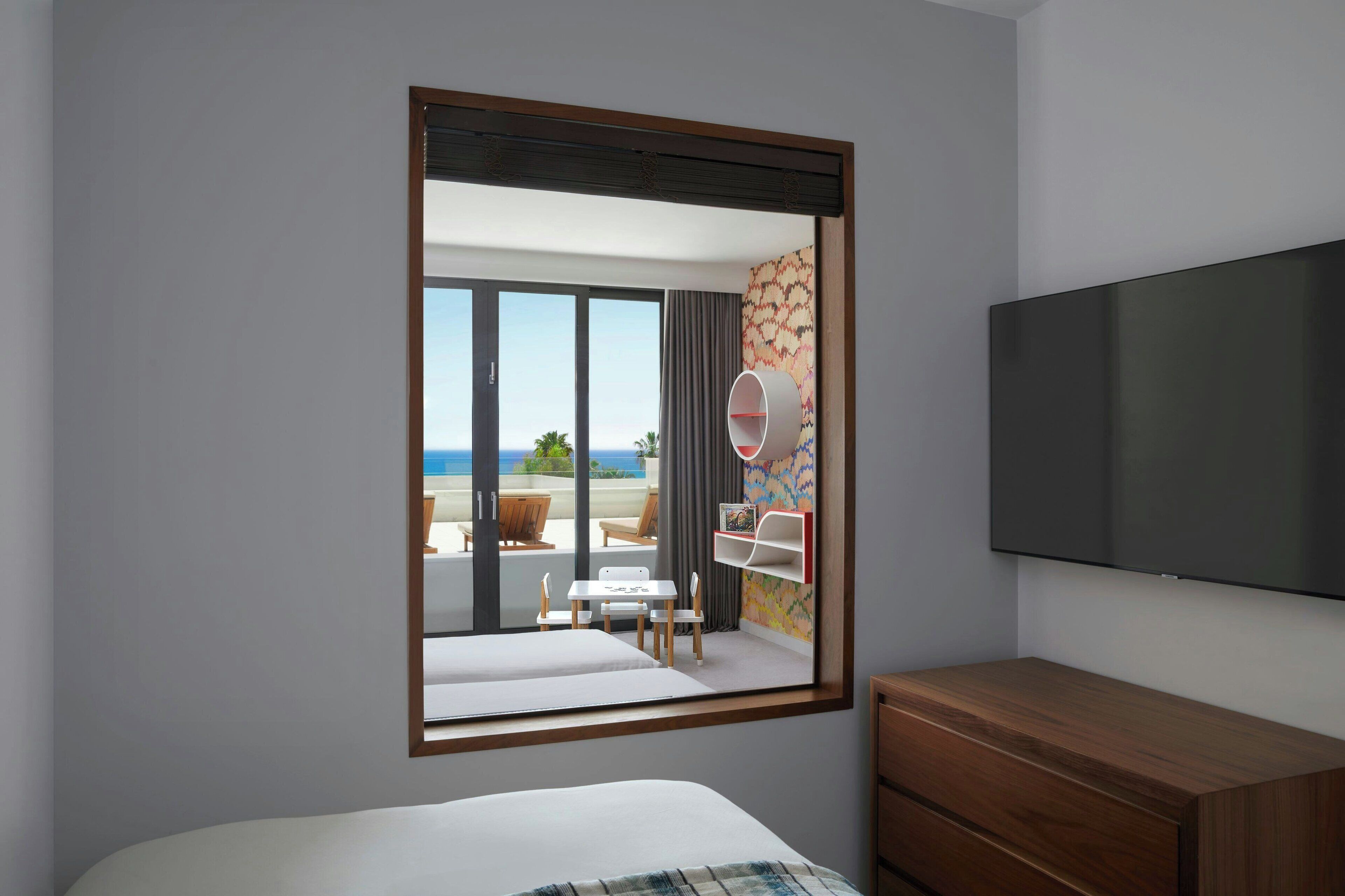 A hotel room with a bed, a wooden dresser, a TV, and a large window with a view of the balcony and outside.