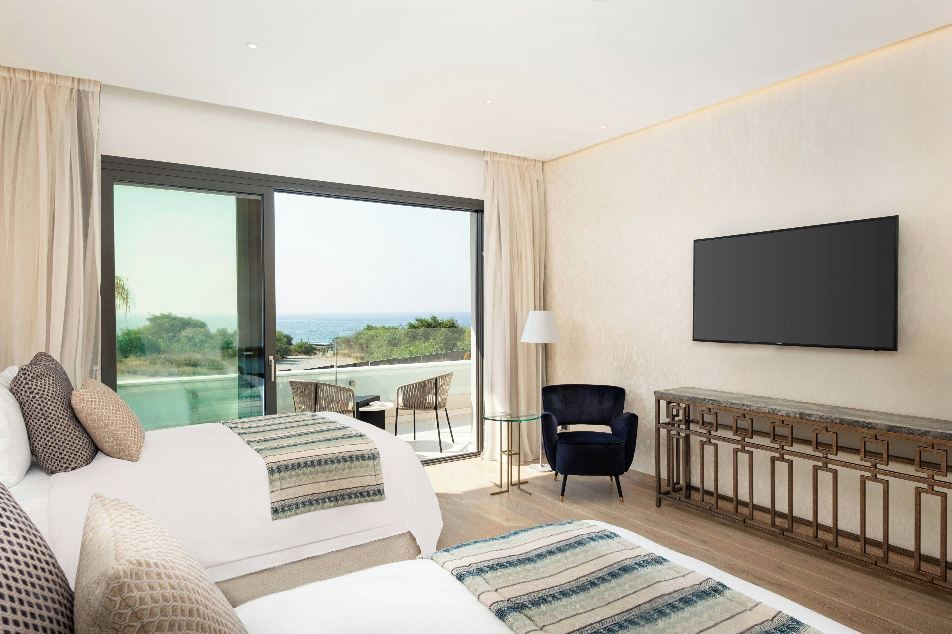 A modern hotel room with a large bed, a TV, a balcony with a table and chairs, and a view of the ocean.