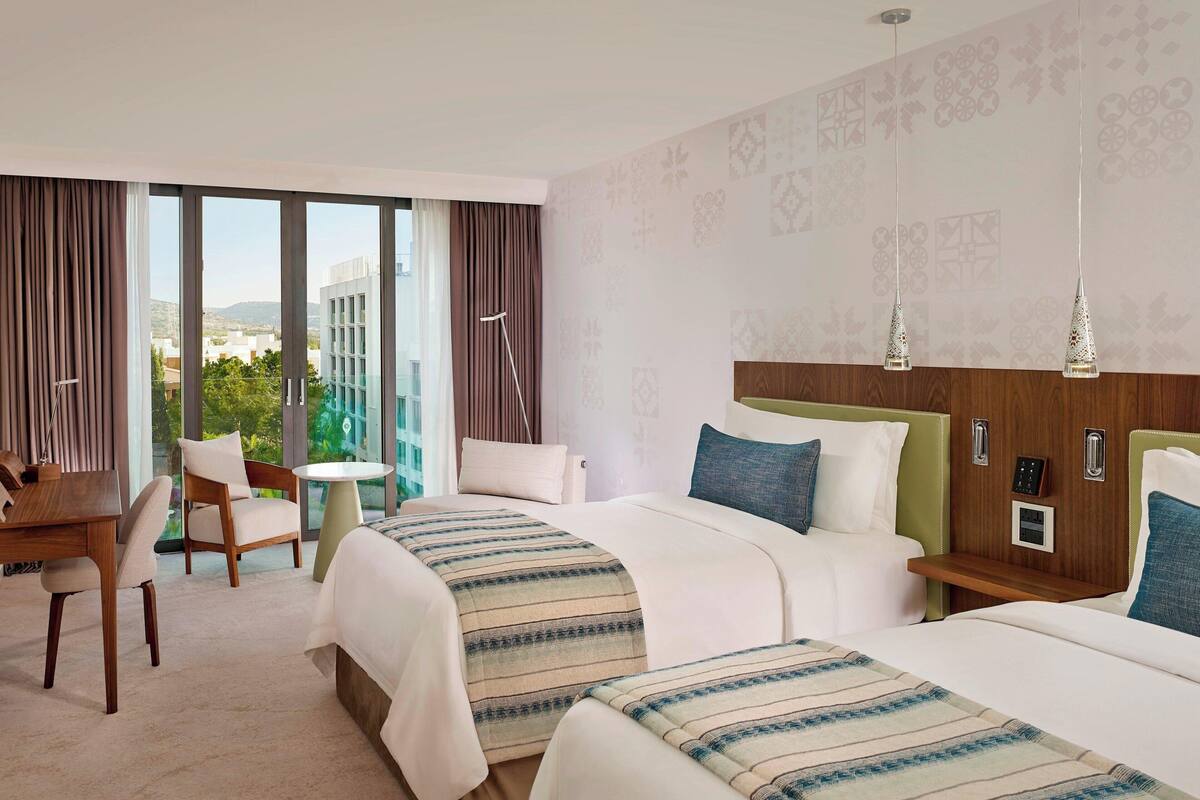 A hotel room with two beds, a desk, a chair, a small table, and a view of the city through large windows.