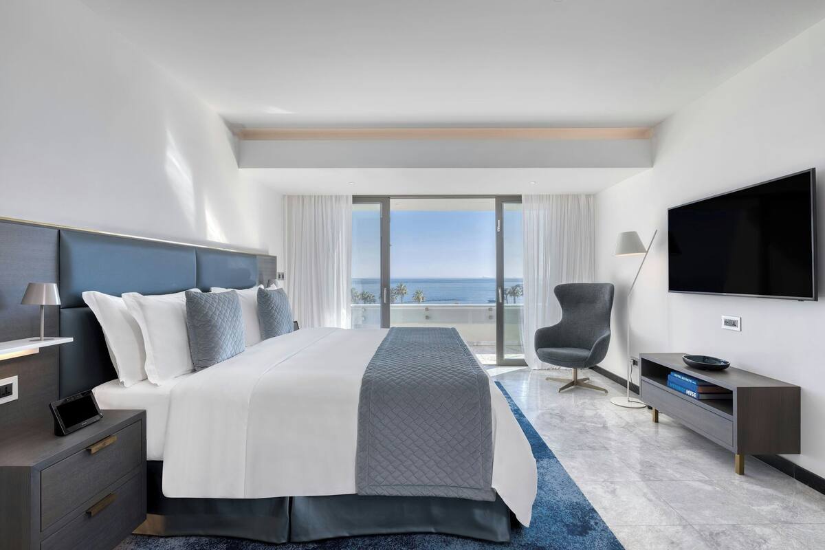 A modern hotel room with a large bed, a TV, a desk, and a view of the ocean.