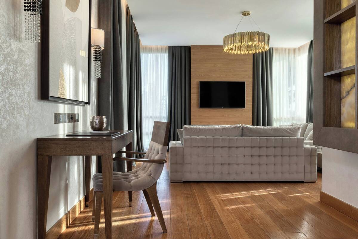 A modern living room with a wooden floor, a sofa, a dining table with chairs, a chandelier, and a flat-screen TV mounted on the wall.