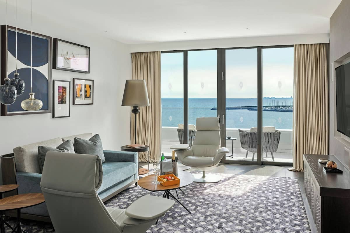 A modern hotel room with a sofa, armchairs, a coffee table, a TV, and a large window with a view of the sea.