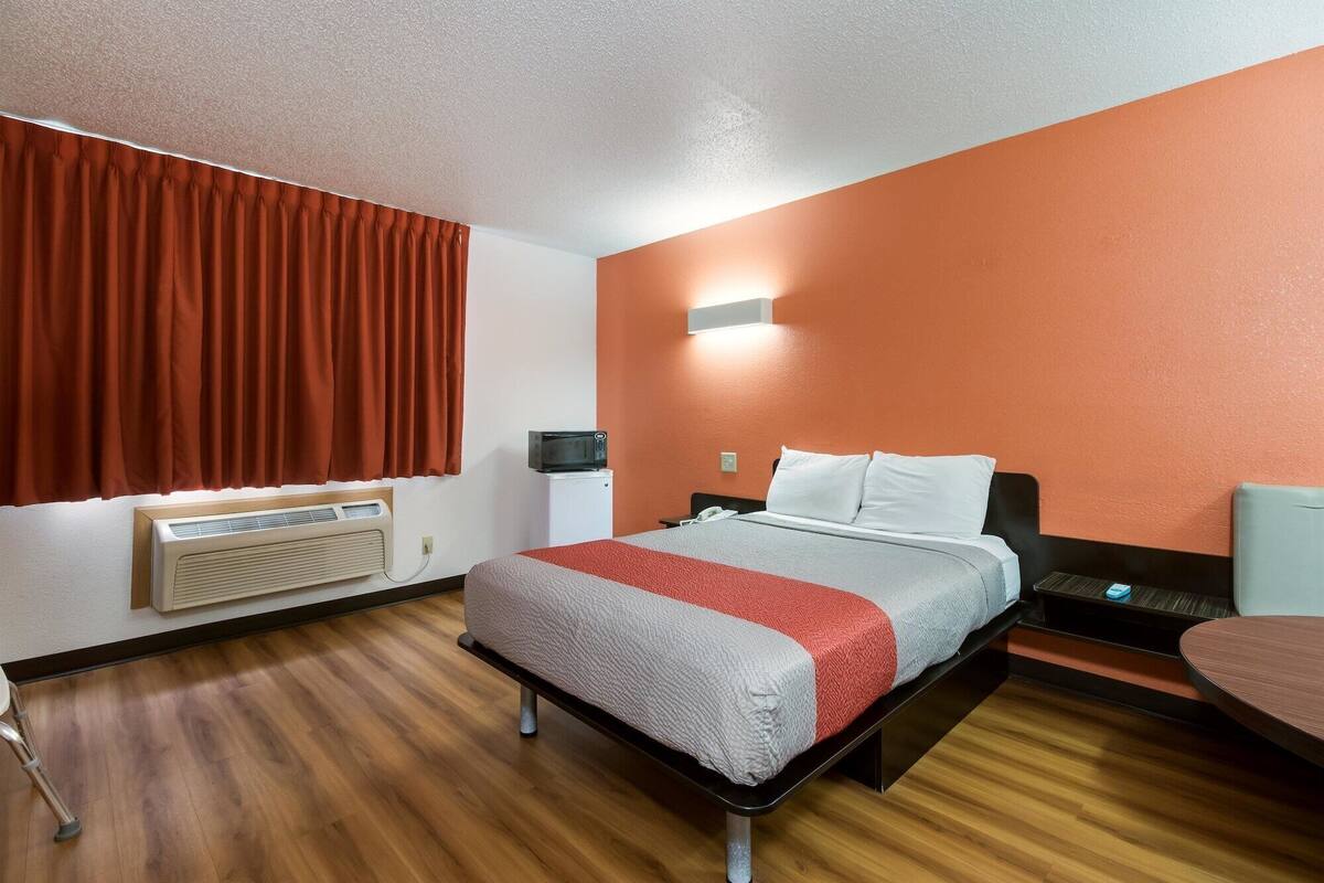 A hotel room with a bed, a bedside table, a chair, a heater, and a window with curtains.