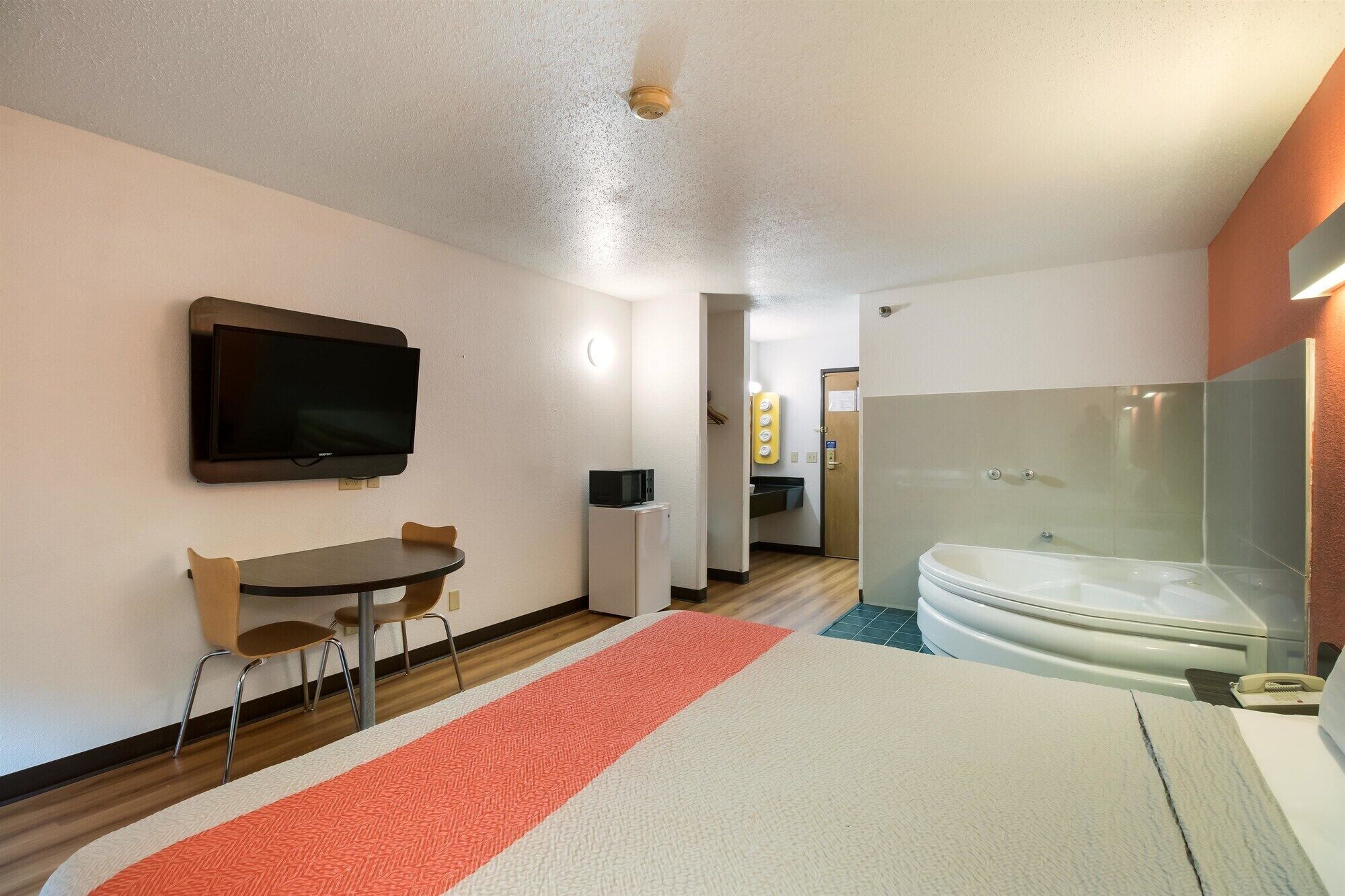 A hotel room with a bed, a TV, a desk, a chair, a microwave, a small refrigerator, and a bathroom with a bathtub and shower.