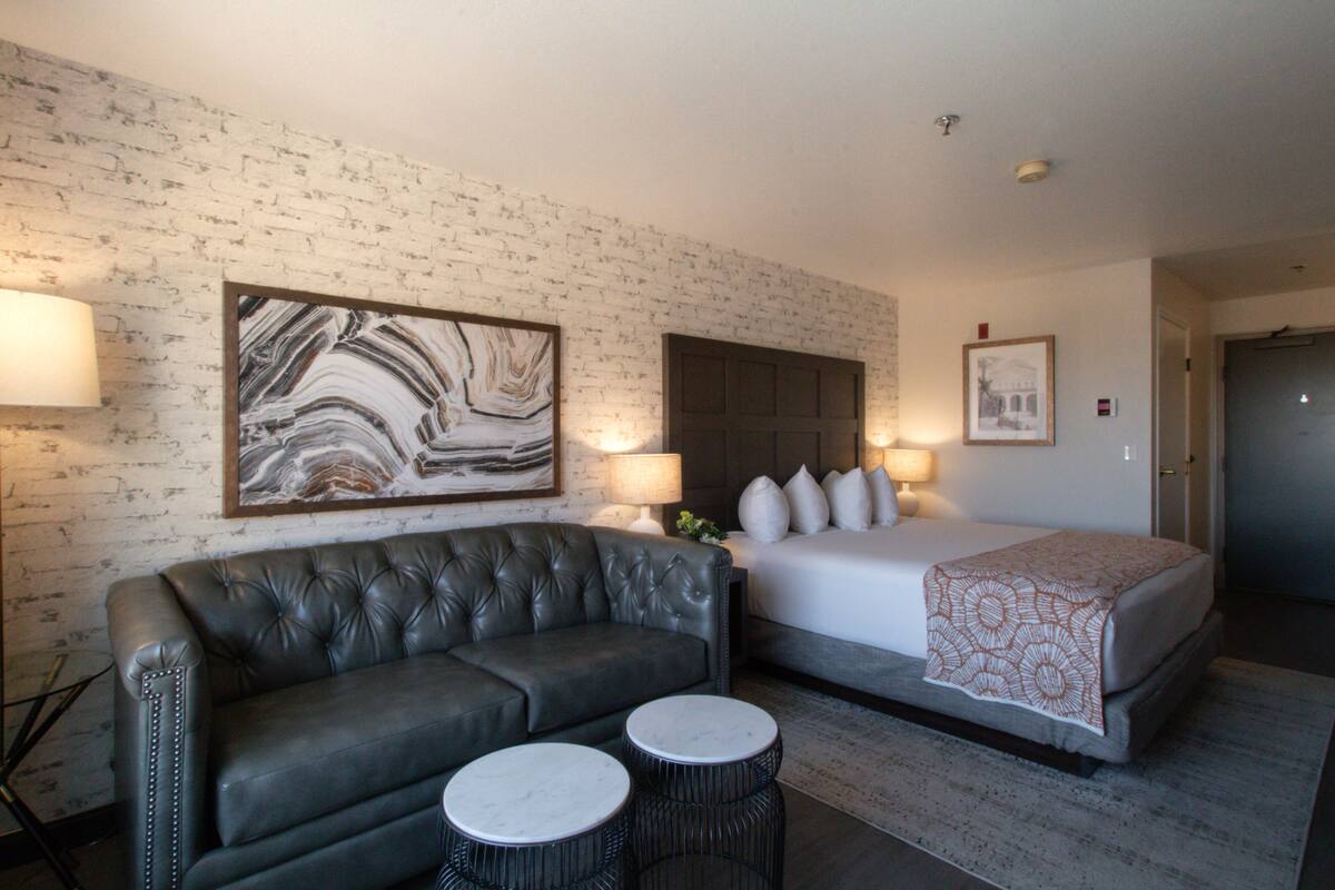 Une chambre d’hôtel avec un canapé en cuir, deux tables rondes, un lit recouvert d’une couverture à motifs et des œuvres d’art accrochées aux murs.