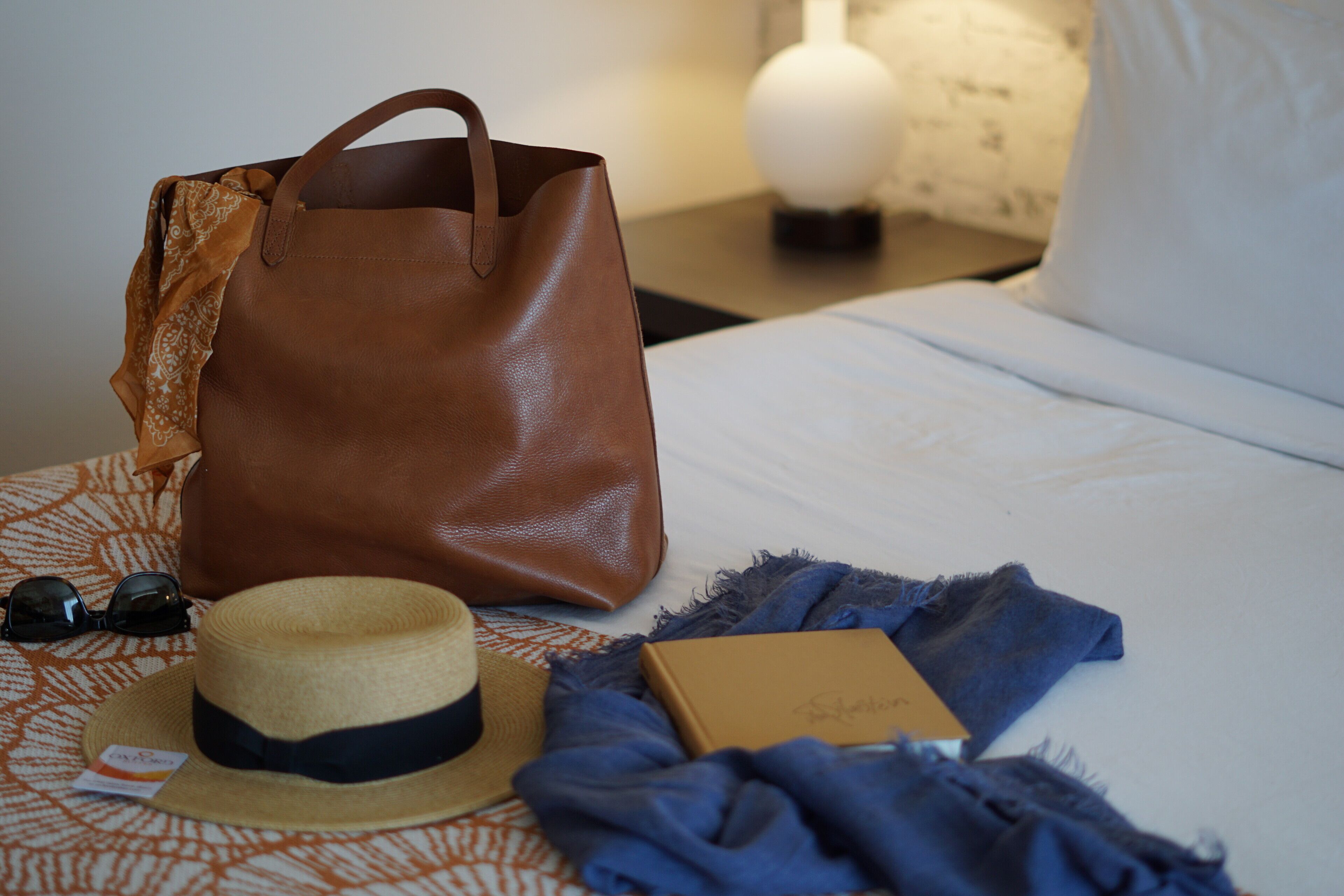 A bed with a brown leather bag, a straw hat, sunglasses, and a box on a patterned bedspread.